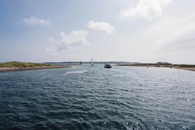 Boats near the channel into Cuttyhunk Harbor between low sandy shorelines.